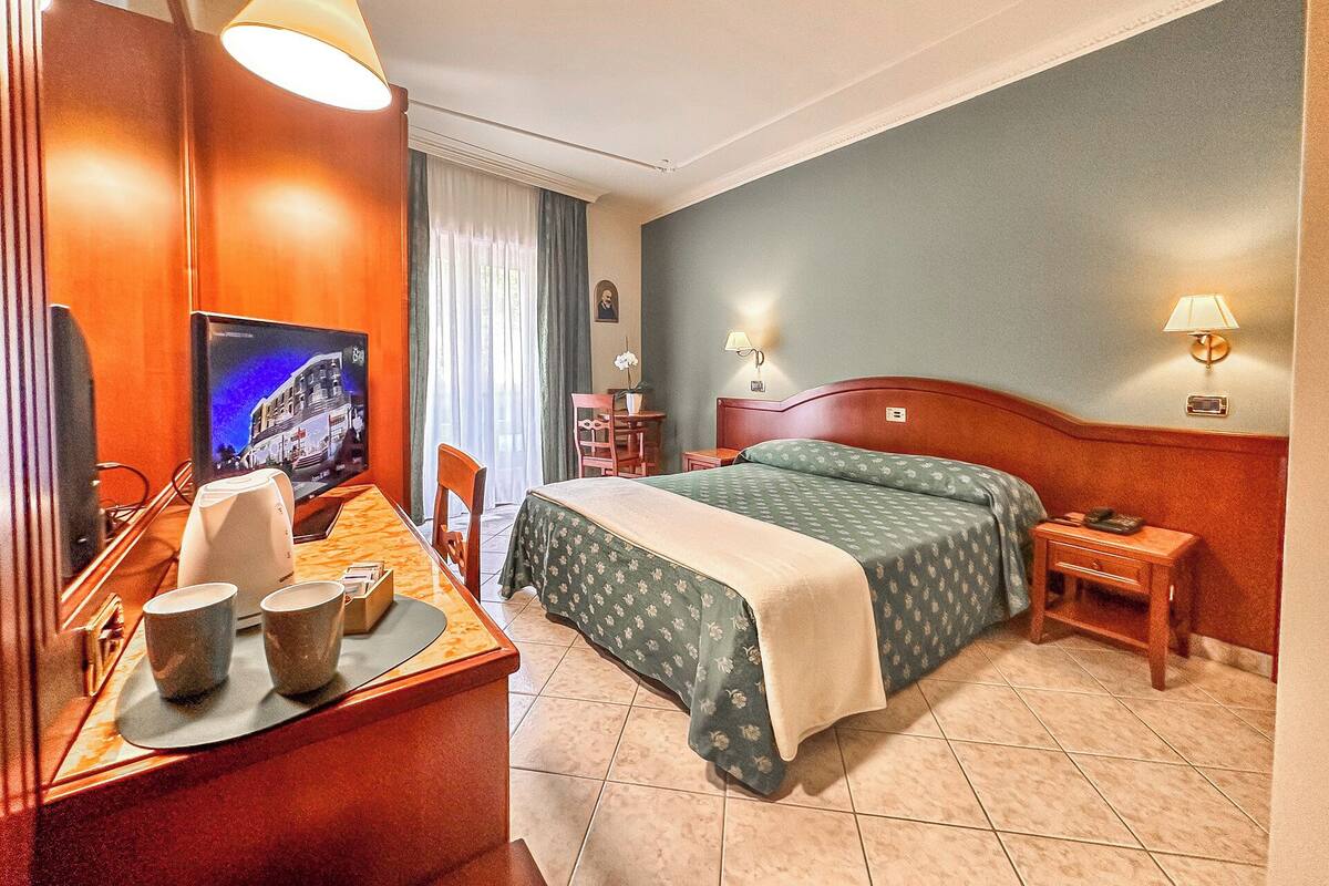A hotel room with a bed, a desk with a television, a chair, and a nightstand with a lamp.