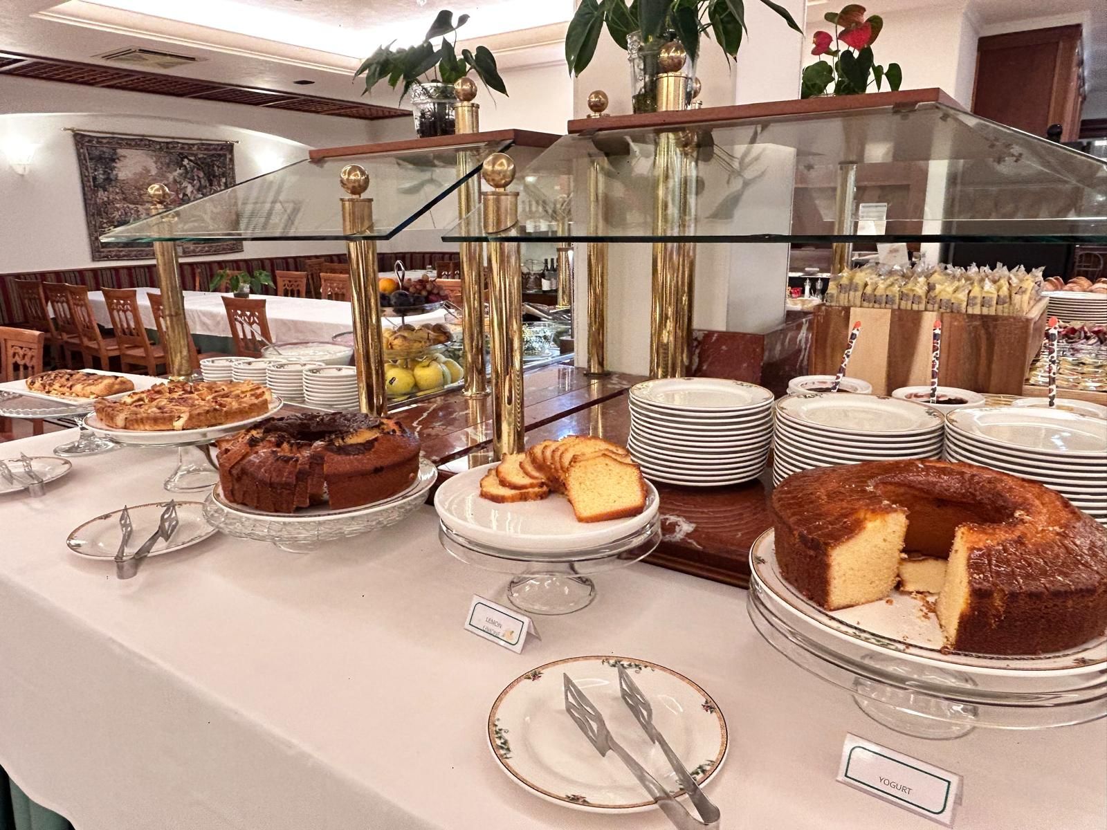 Un bufé con diversos alimentos, entre ellos pasteles y productos de pastelería, y una vitrina con platos y cubiertos.