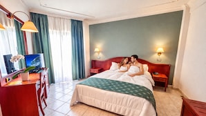 Een hotelkamer met een bed, nachtlampjes, een bureau met een computer, een televisie en een potplant.