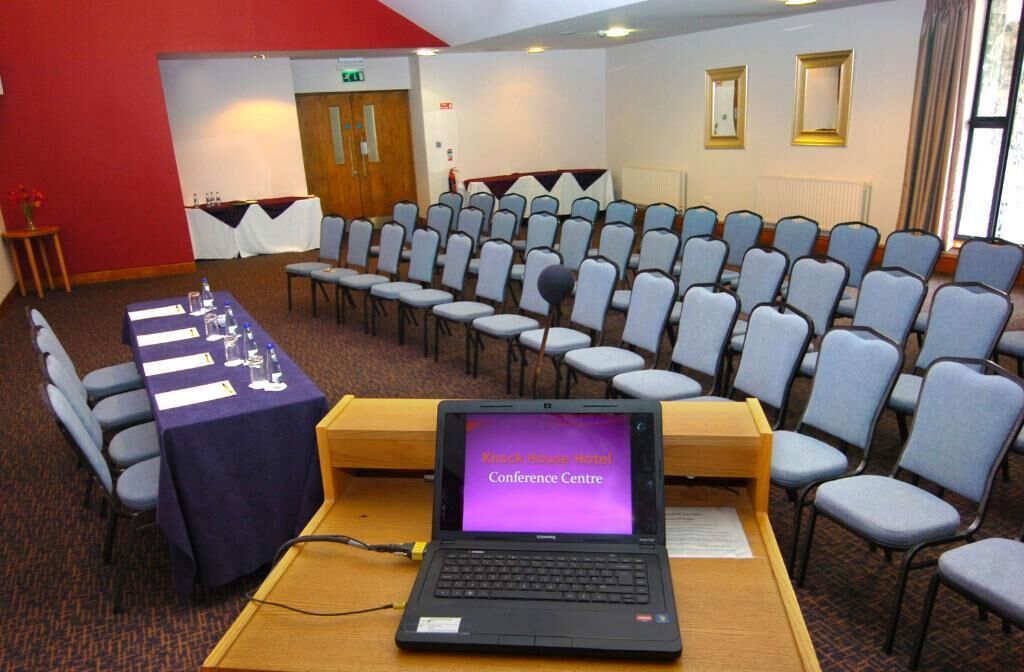 A conference room with rows of chairs, a table with water bottles, and a laptop displaying 'Knock Knock Hotel Conference Centre'.