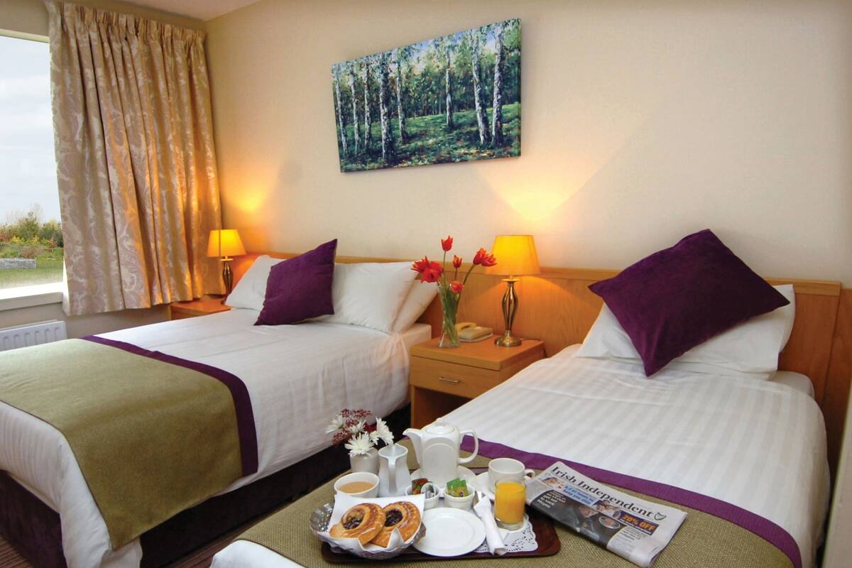 A hotel room with two beds, a painting of a forest, a window with curtains, and a breakfast tray on the floor.