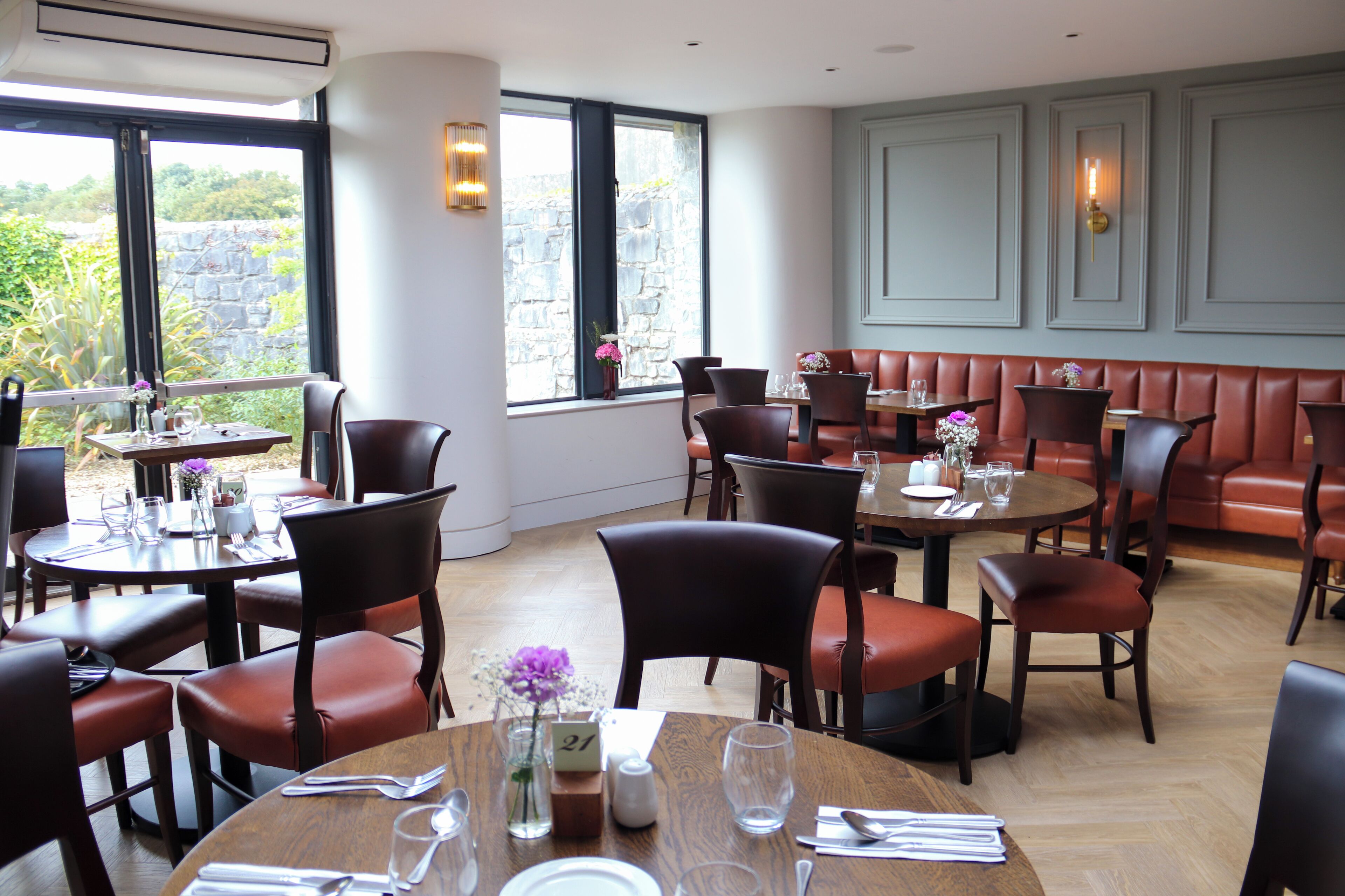 A restaurant with round tables and booths, large windows with a stone wall view, and a light-colored wooden floor.