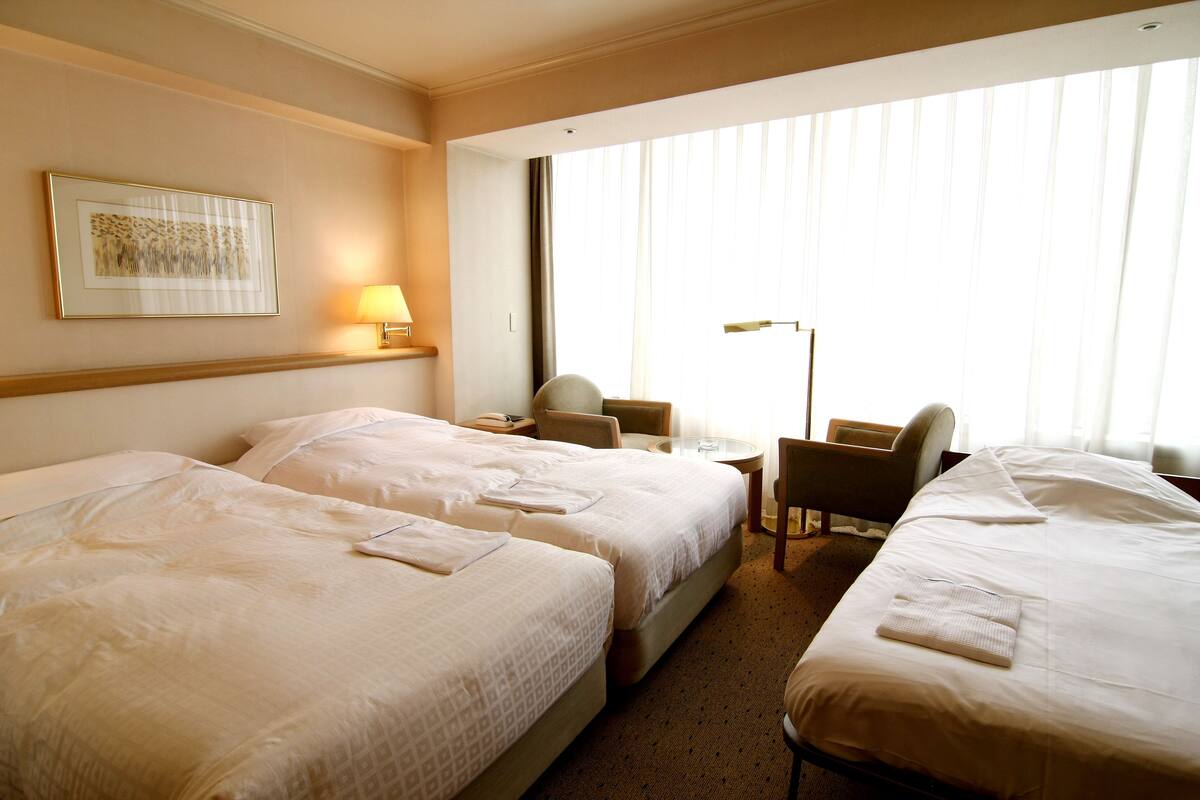 Une chambre d’hôtel avec deux lits, un bureau, une chaise et une grande fenêtre avec des rideaux.