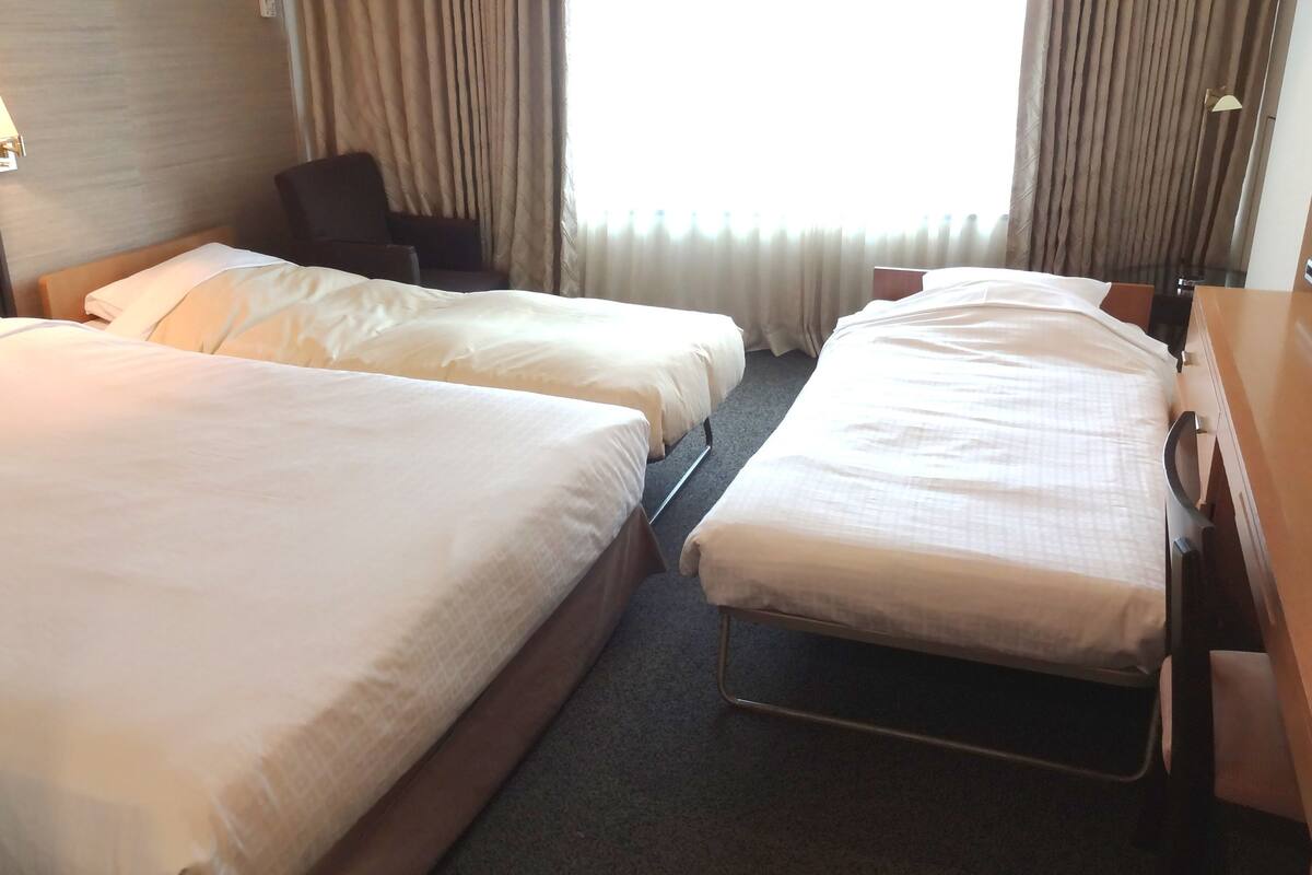 Une chambre d’hôtel avec deux lits simples, un bureau avec une chaise, une lampe et une fenêtre avec des rideaux.