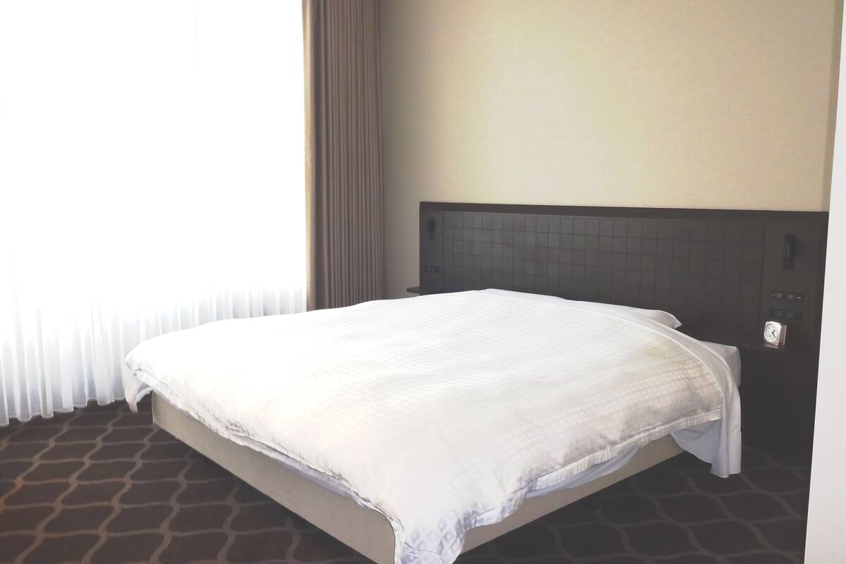 Une chambre d’hôtel avec un lit, une chaise, une fenêtre avec des rideaux et une horloge au mur.