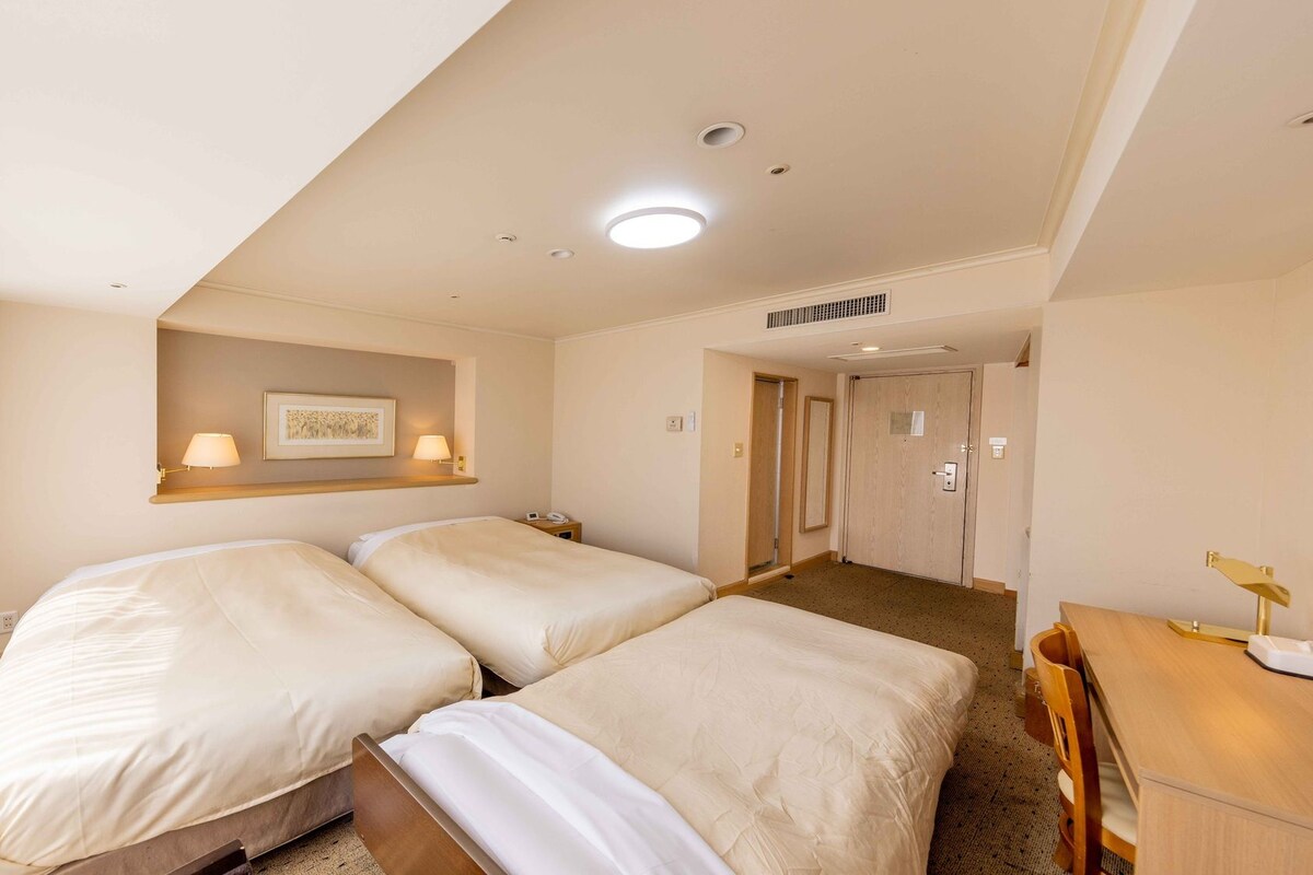 A hotel room with two beds, a desk, a chair, a framed picture on the wall, and a door leading to another room.