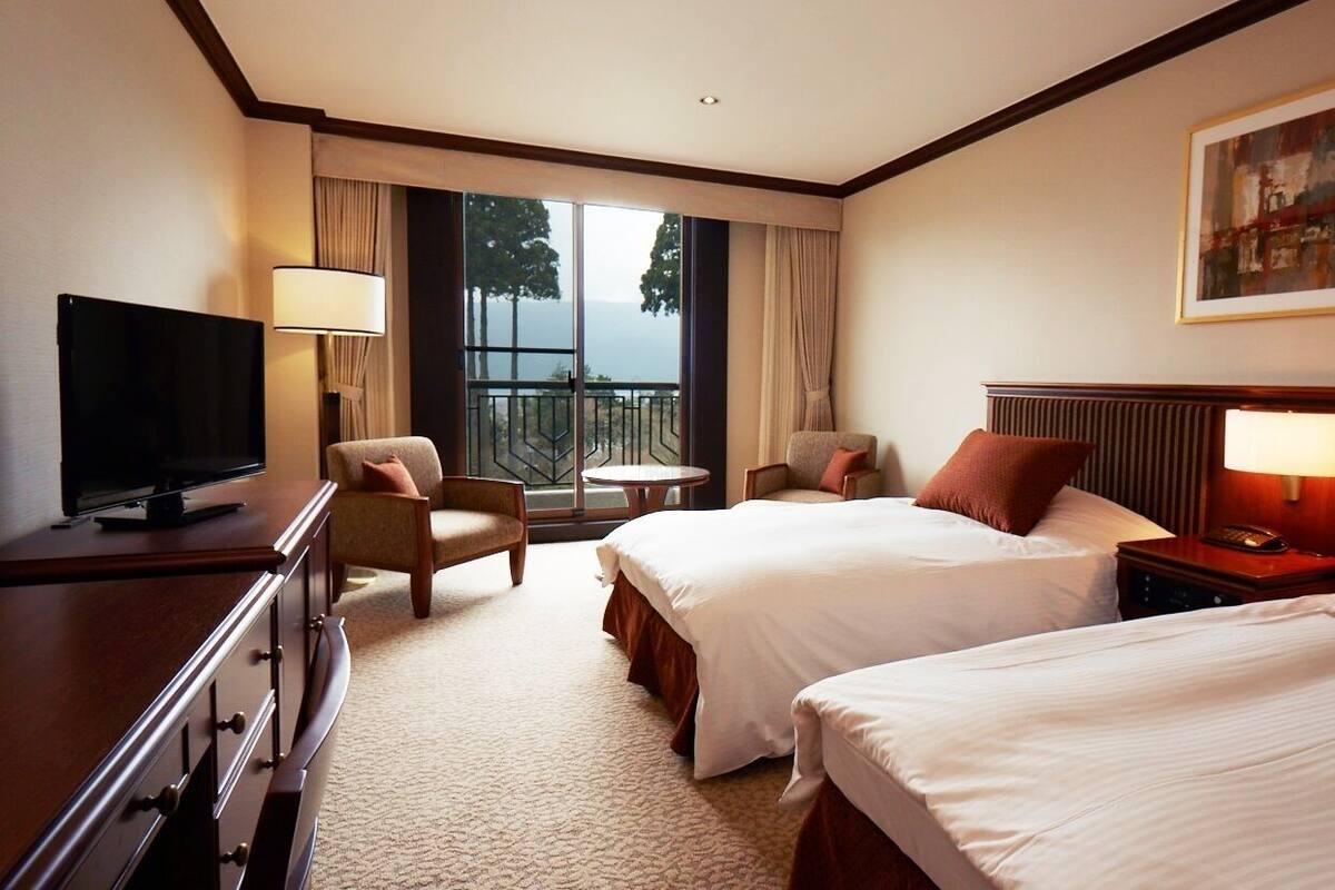 A hotel room with two beds, a TV, a desk, and a balcony with a view.