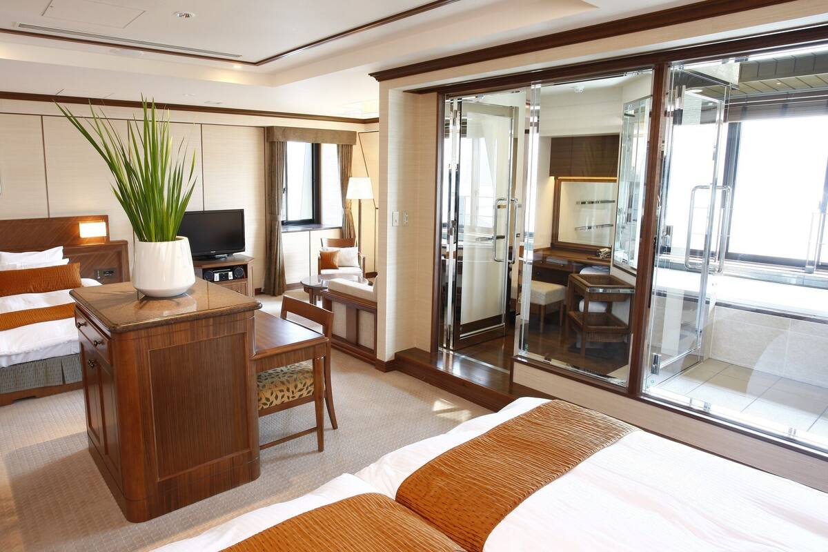 A hotel room with a bed, a desk with a plant, a TV, a seating area, and a sliding glass door to a balcony.