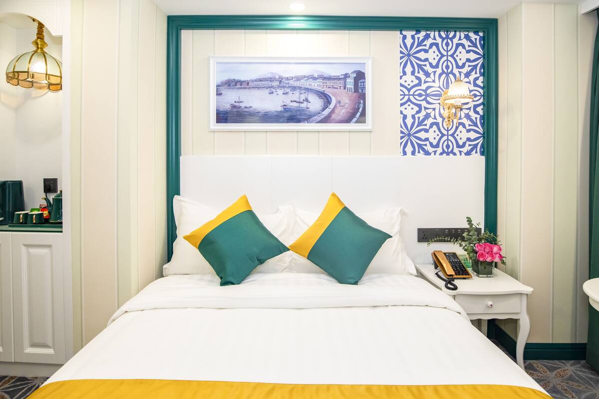A hotel room with a bed, a bedside table with a phone and flowers, and a wall with a painting and patterned wallpaper.