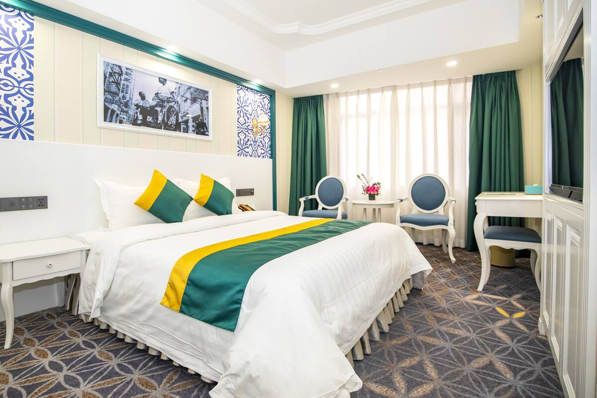 A hotel room with a large bed, a desk, two chairs, a vanity with a mirror, and a window with curtains.