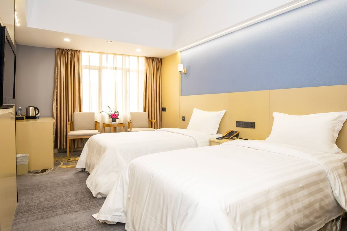 A hotel room with two beds, a desk, a chair, a small table with a flower vase, and a window with curtains.