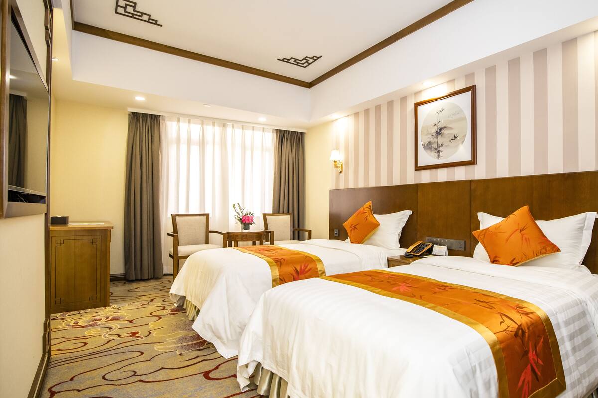 A hotel room with two beds, a desk, a chair, a small table with a vase, and a painting on the wall.