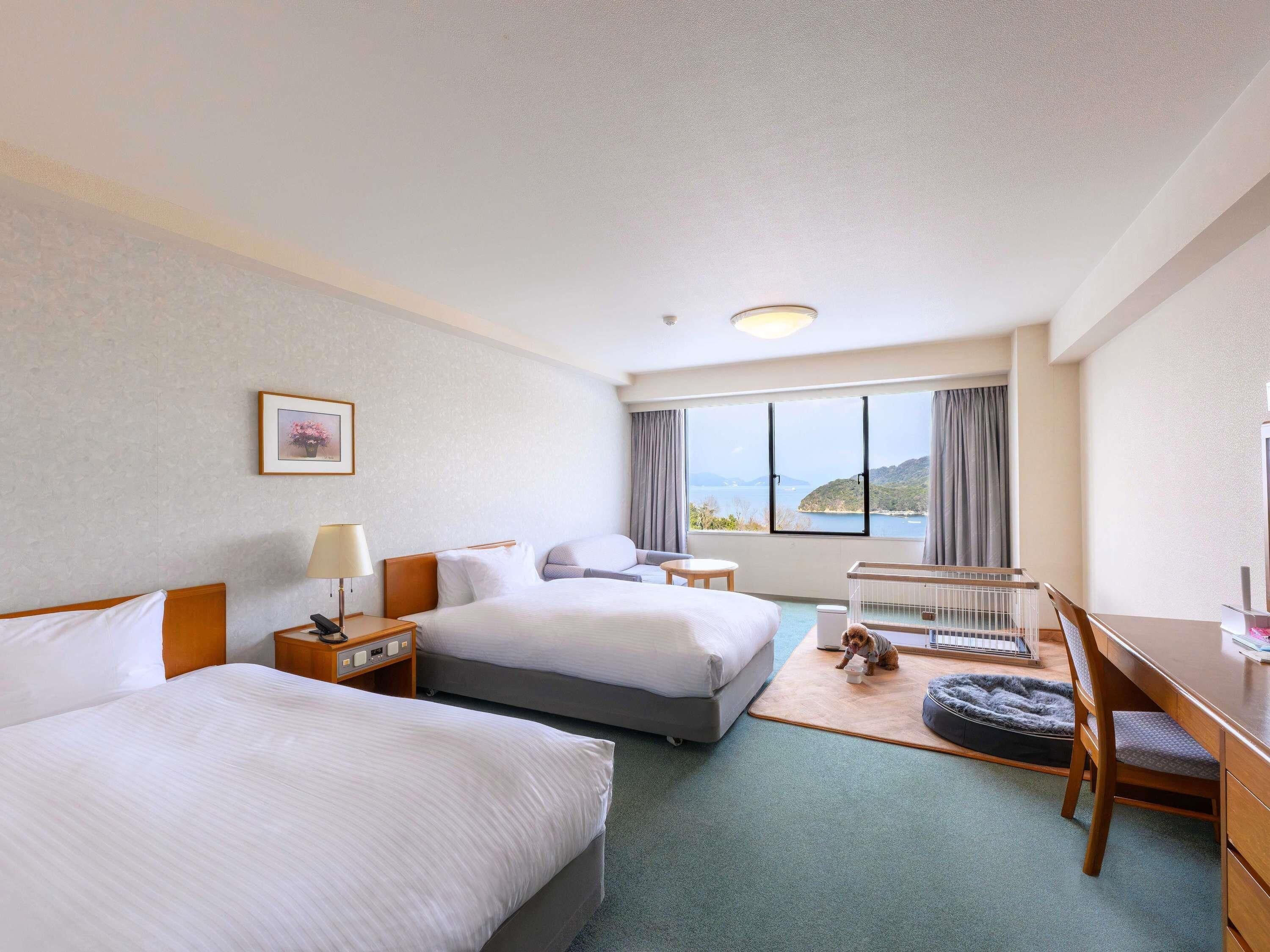 Habitación de hotel con dos camas, un escritorio, una silla, una cama para mascotas y un ventanal con vistas.