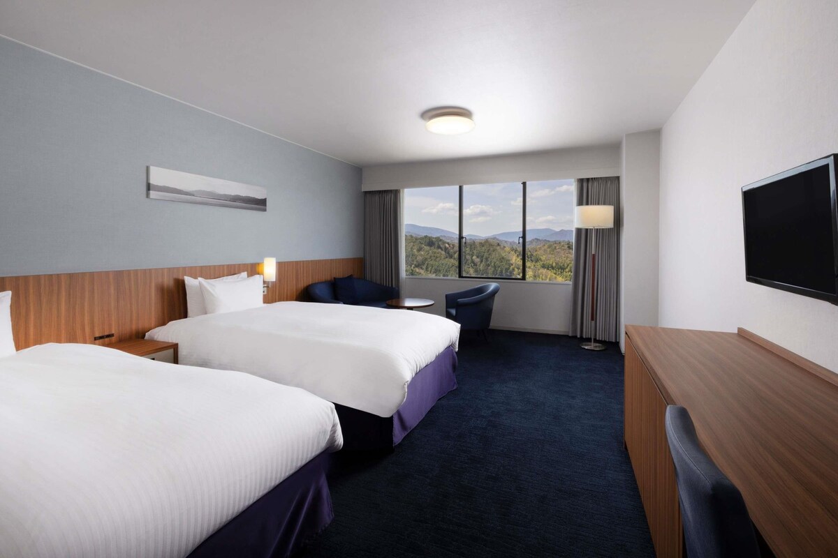 A hotel room with two beds, a desk, a TV, and a large window with a mountain view.
