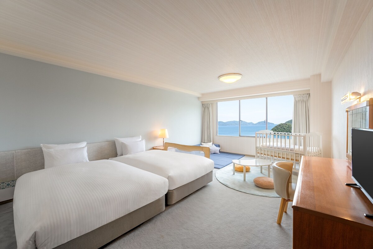 Ein Hotelzimmer mit zwei Betten, einem Schreibtisch, einem Fernseher und einem Balkon mit Meerblick.