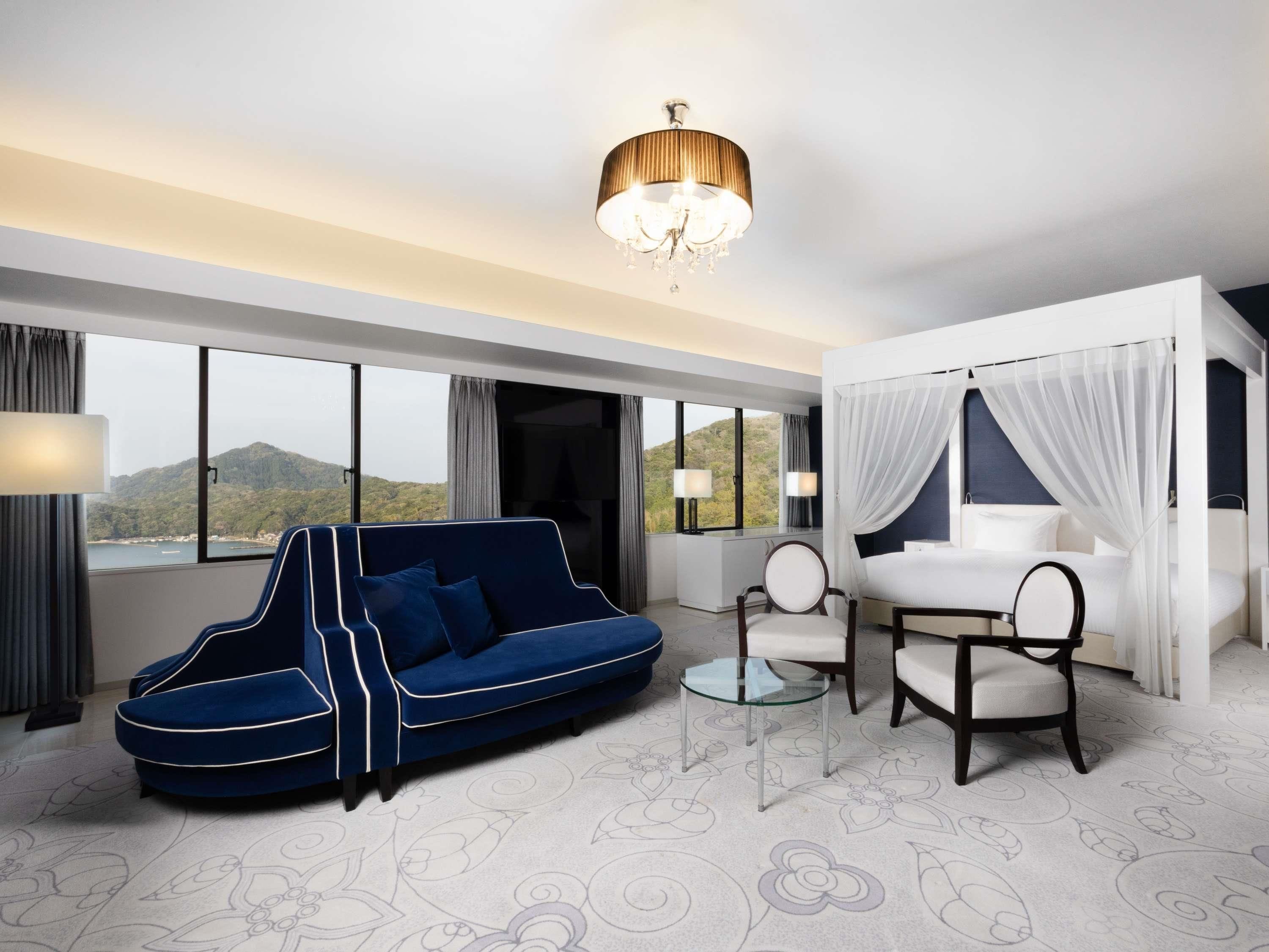 A modern hotel room with a large window, a blue sofa, a white bed with a canopy, and a chandelier.