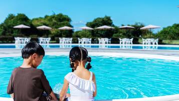 Dos niños sentados junto a una piscina, con sombrillas y sillas al fondo.