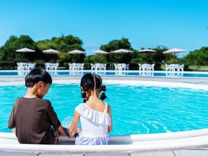 Dos niños sentados junto a una piscina, con sombrillas y sillas al fondo.