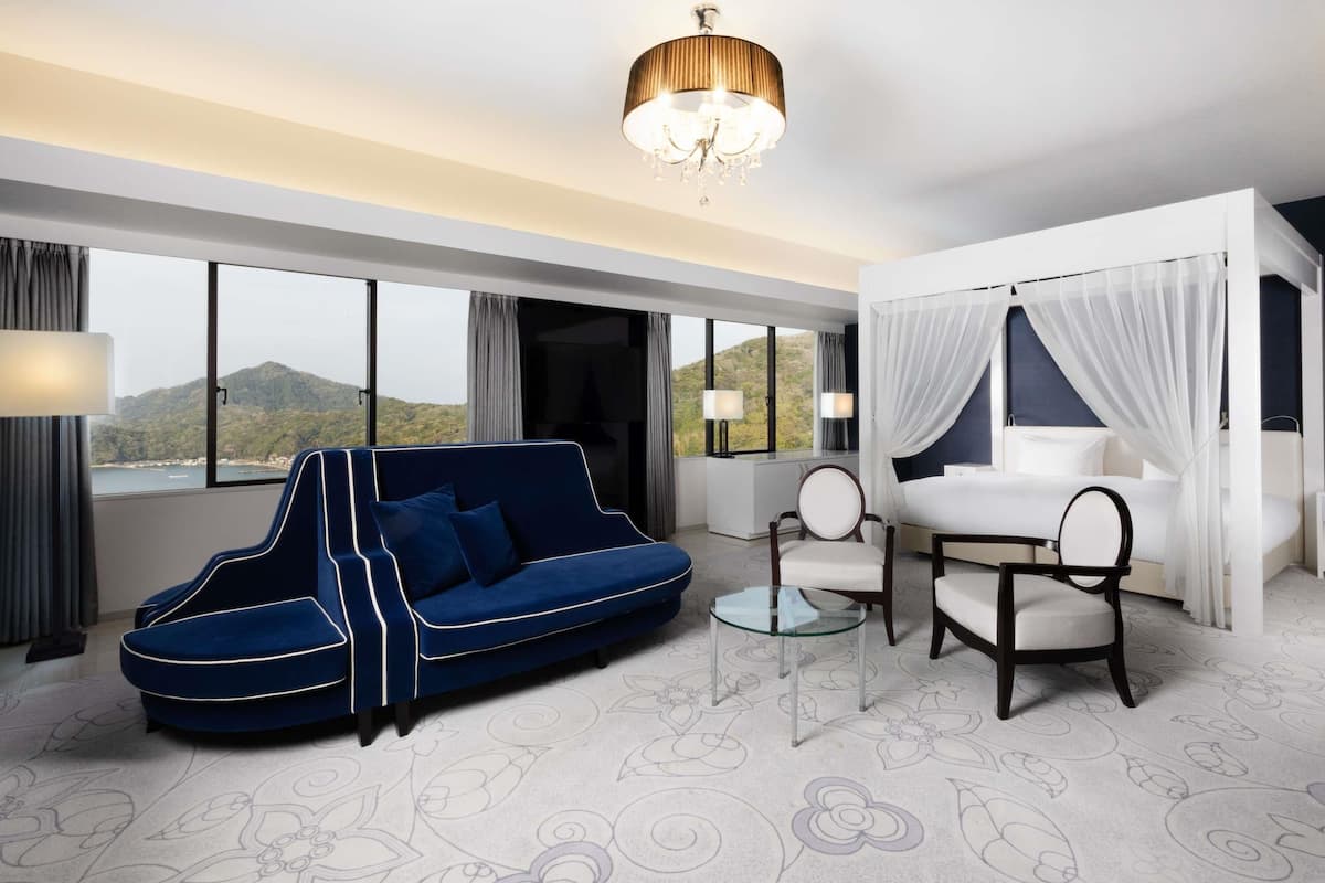 A modern hotel room with a large window, a blue sofa, a white bed with a canopy, and a chandelier.