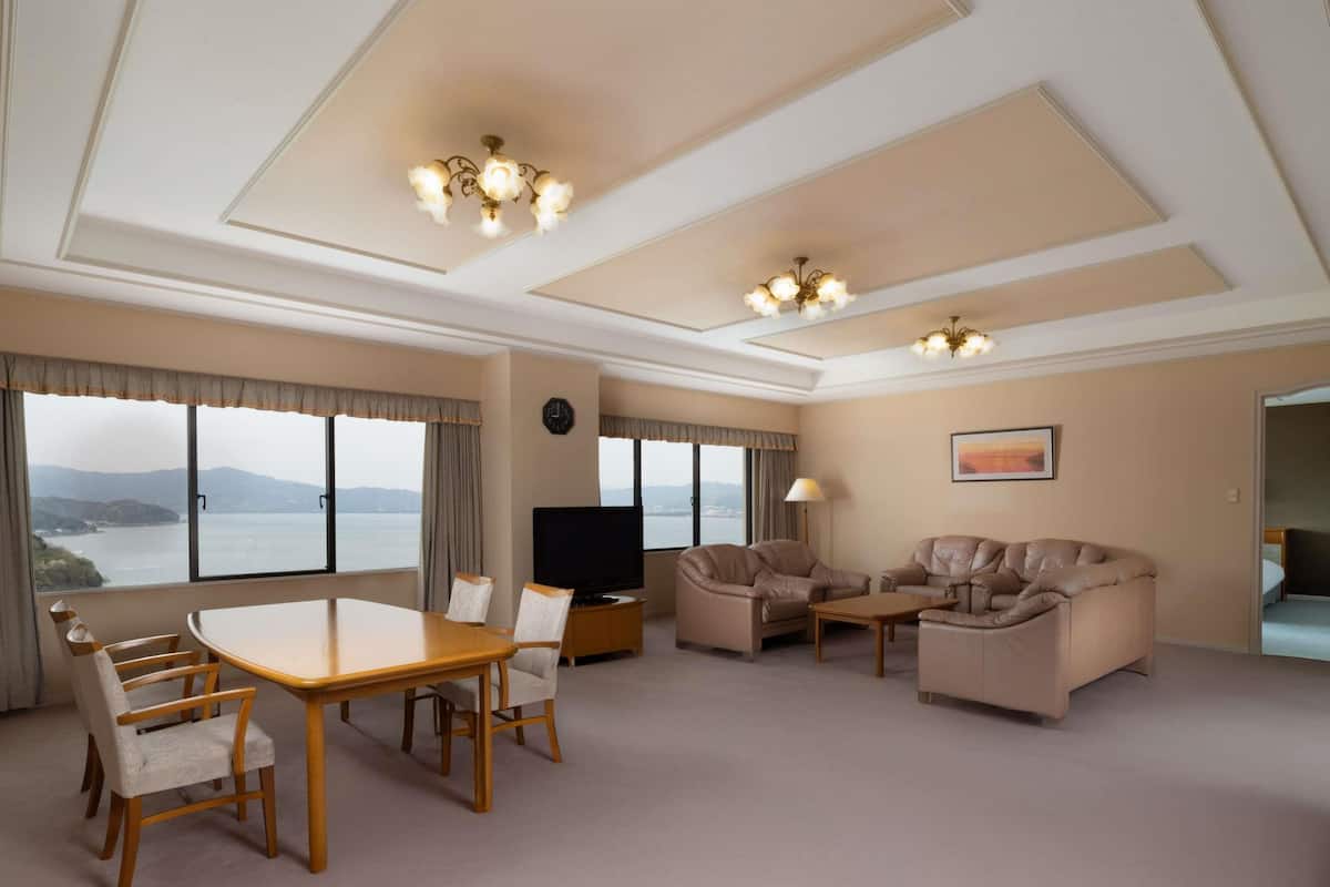 A spacious hotel room with a dining area, two sofas, a TV, and large windows offering a view of the sea.