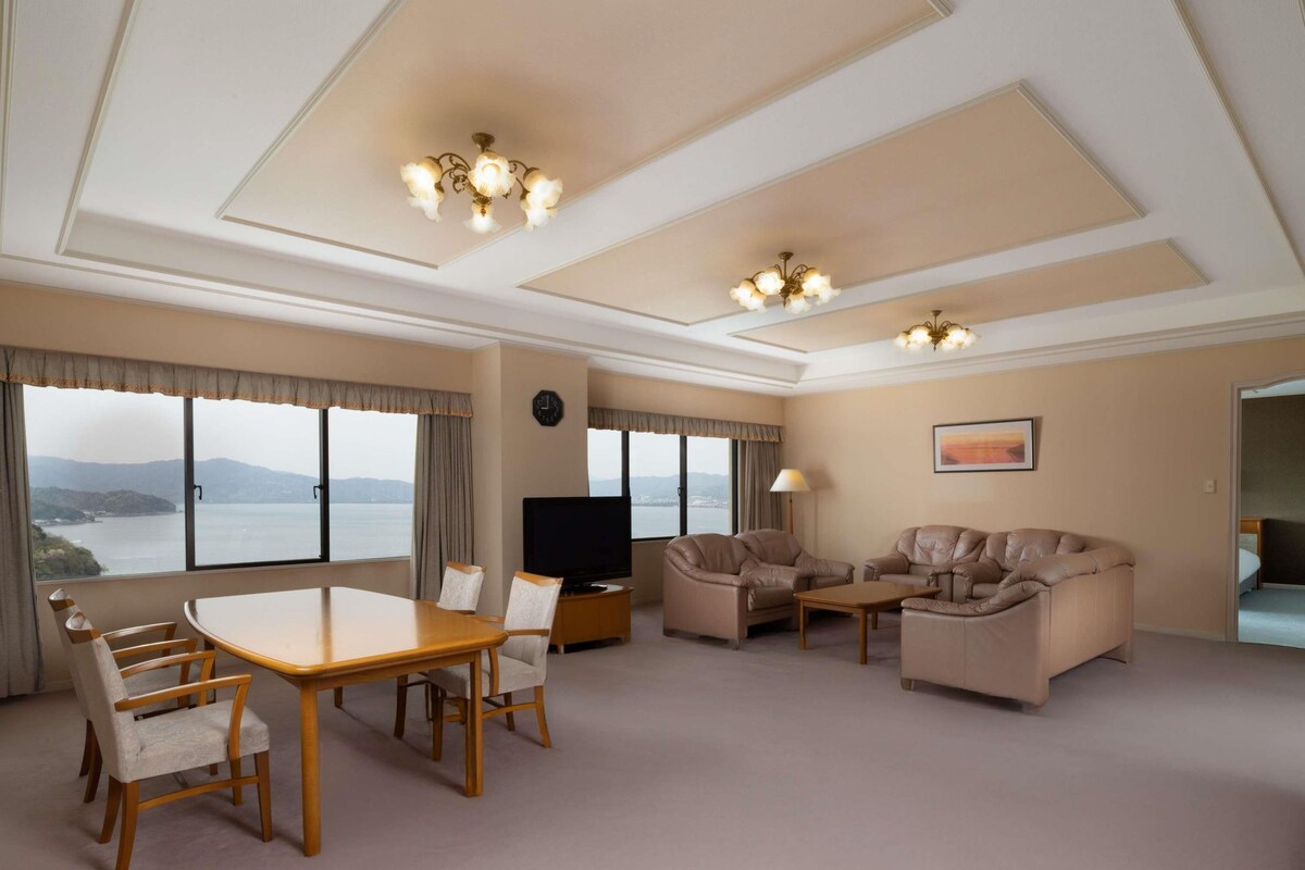 A spacious hotel room with a dining area, two sofas, a TV, and large windows offering a view of the sea.