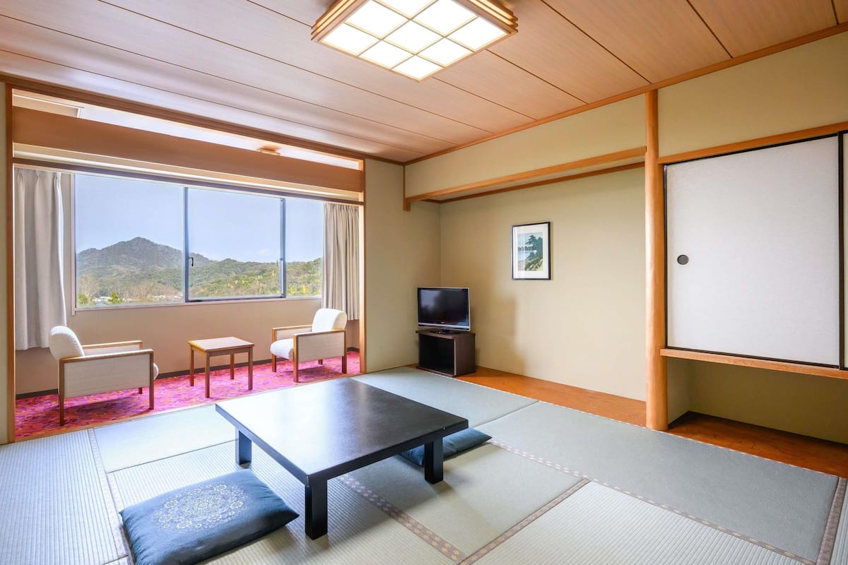 A room with tatami flooring, a low table, two chairs, a television, and a window with a view of hills.