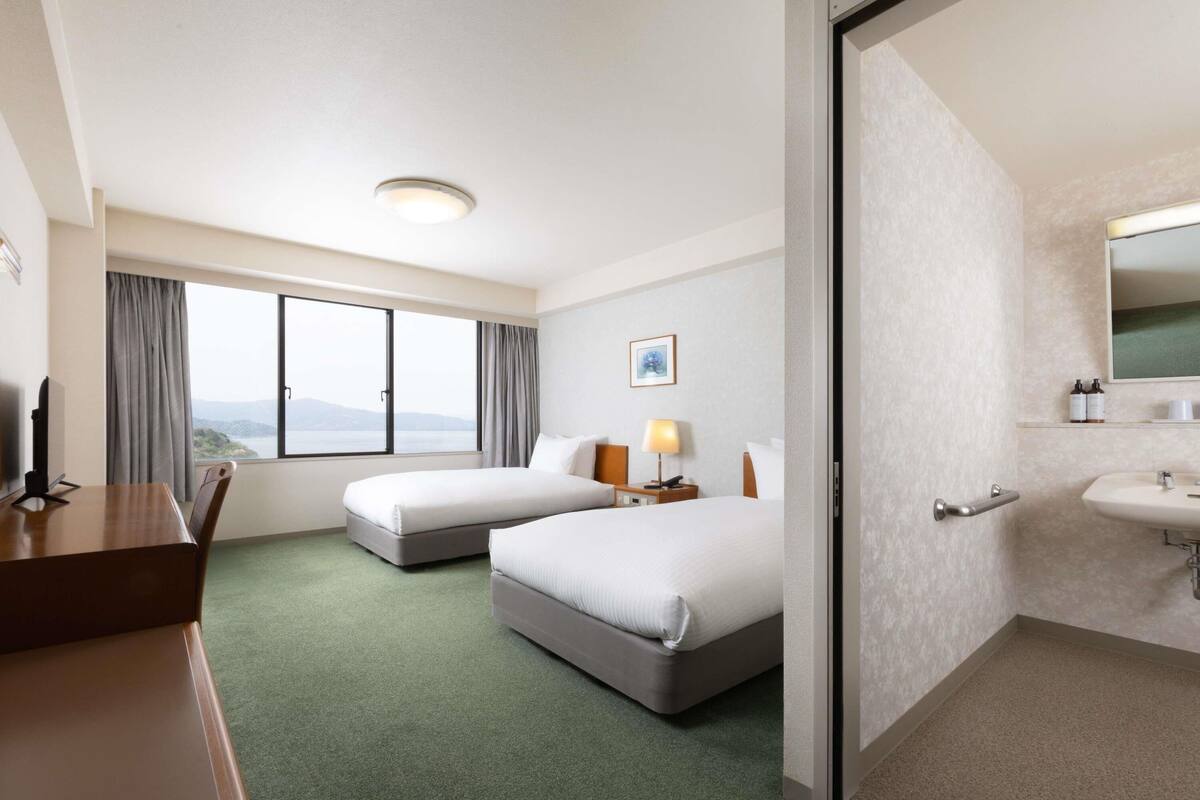 A hotel room with two beds, a desk, a chair, a window with a view, a bedside lamp, and a bathroom with a sink and mirror.