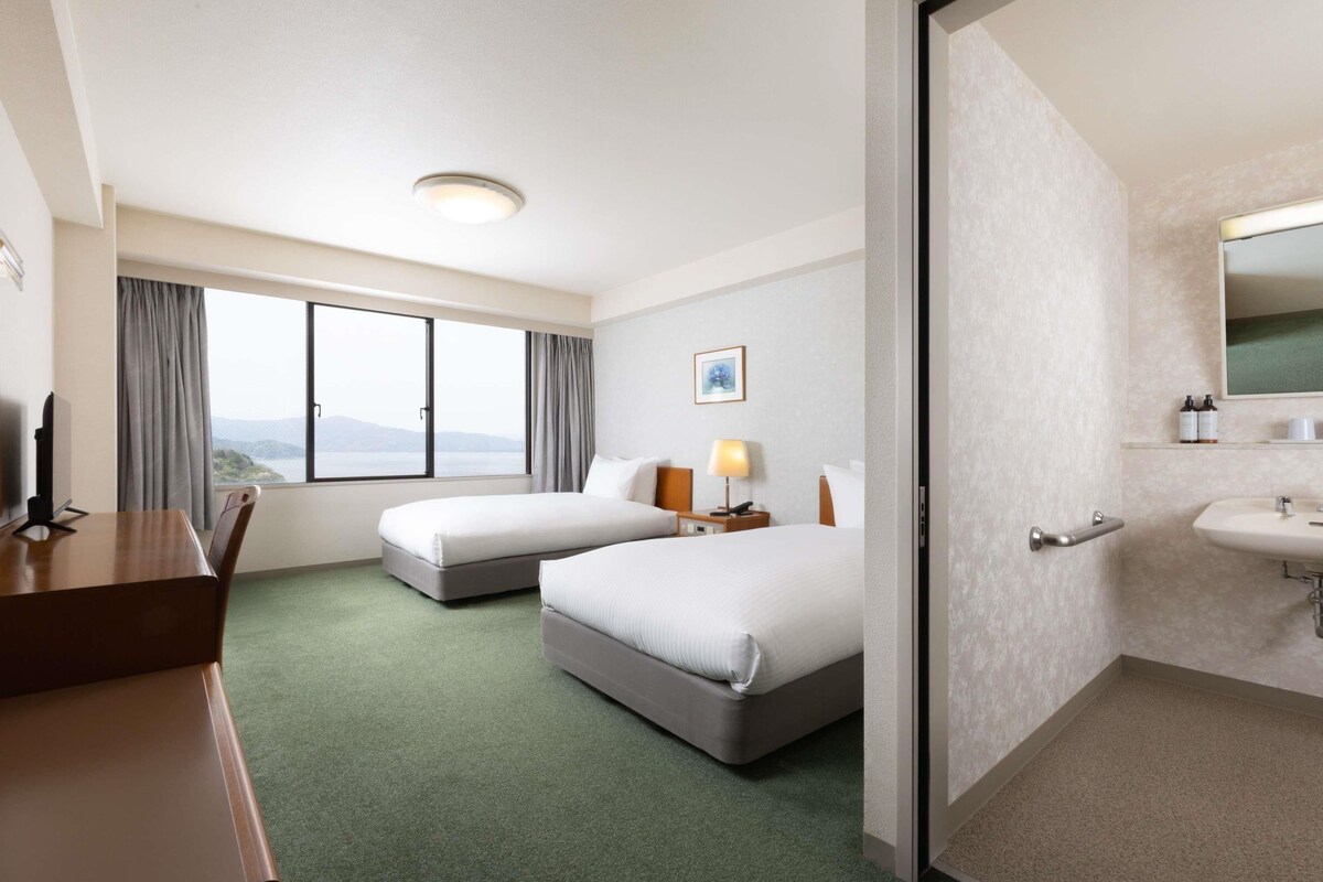 A hotel room with two beds, a desk, a chair, a window with a view, a bedside lamp, and a bathroom with a sink and mirror.