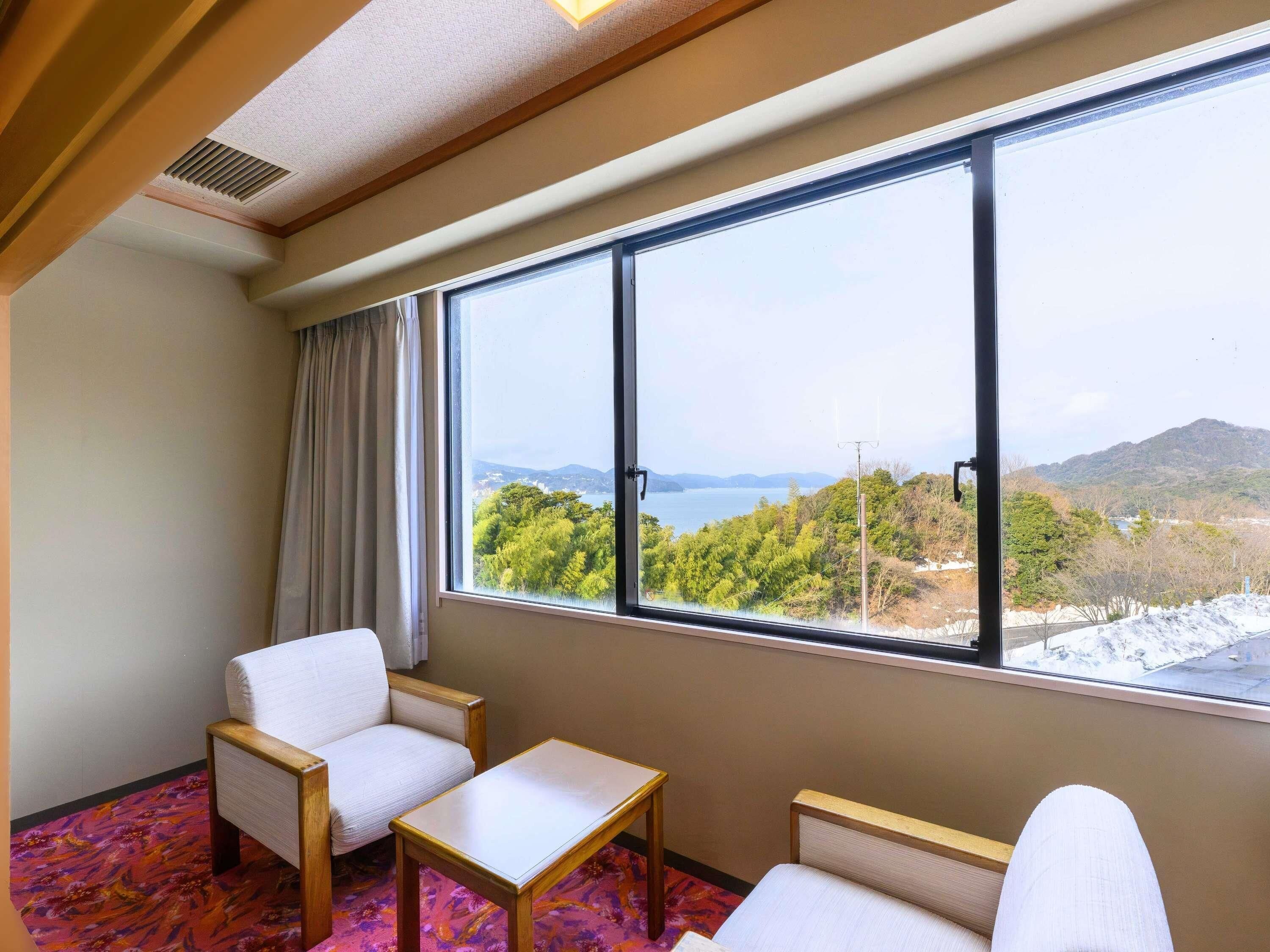 A hotel room with a large window, two white chairs, a small table, and a patterned carpet.