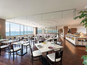 Um restaurante amplo com várias mesas e cadeiras, janelas grandes com vista para o mar e um buffet com diversos alimentos.