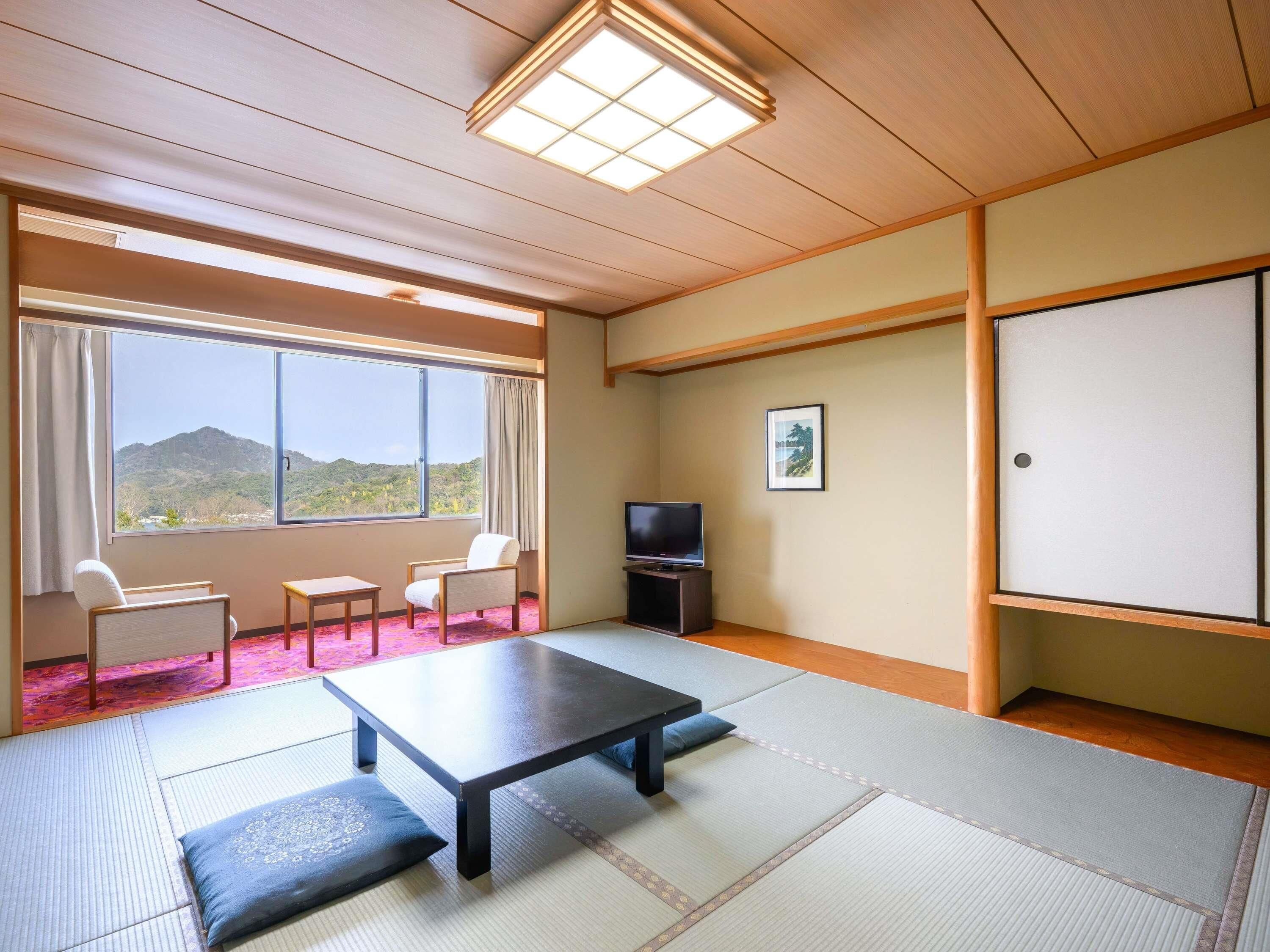 Een kamer met tatami vloerbedekking, een lage tafel, twee stoelen, een televisie en een raam met uitzicht op heuvels.
