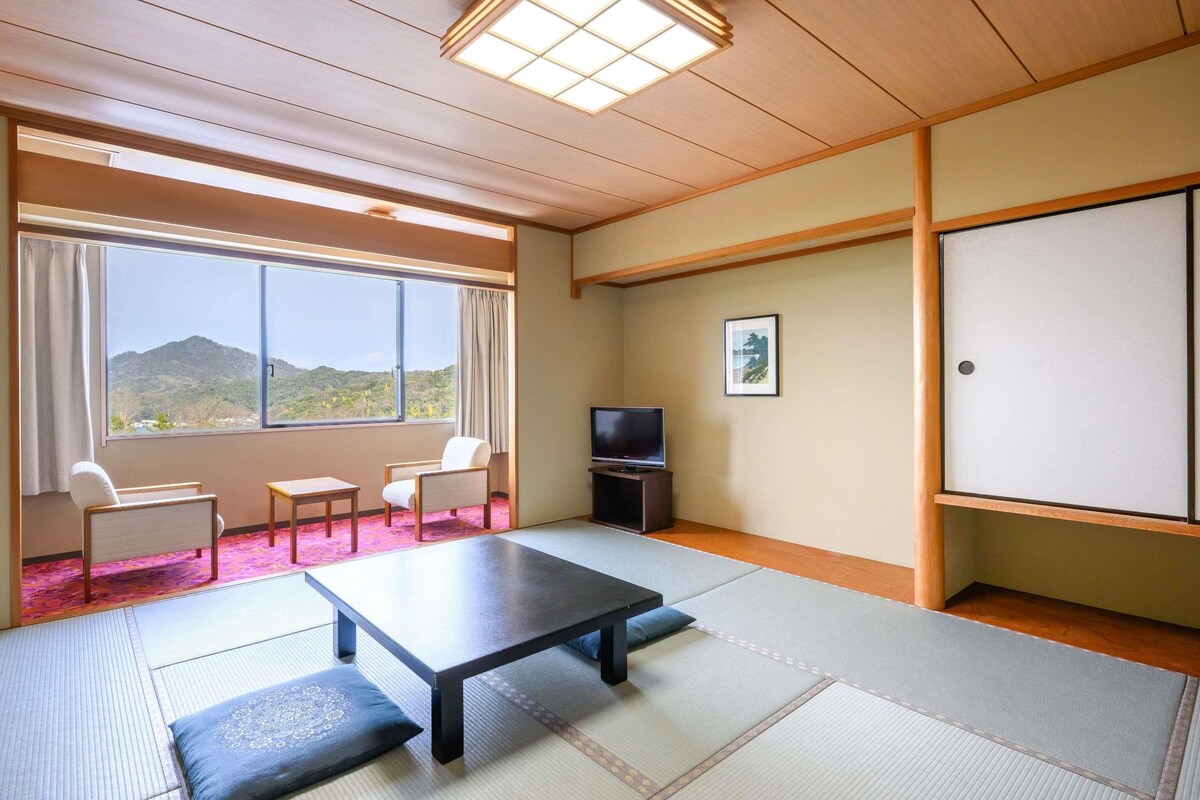 A room with tatami flooring, a low table, two chairs, a television, and a window with a view of hills.