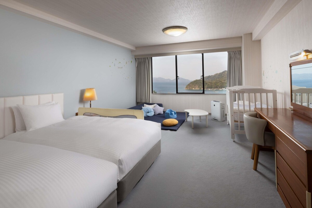A hotel room with a large bed, a desk, a chair, a small table, and a view of the sea.