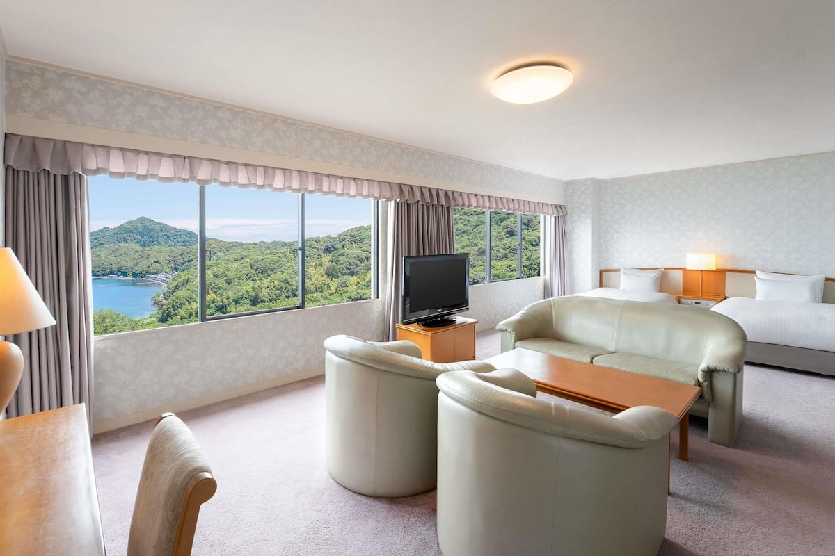 A hotel room with a large window, a flat-screen TV, a sofa, a chair, and a small table.