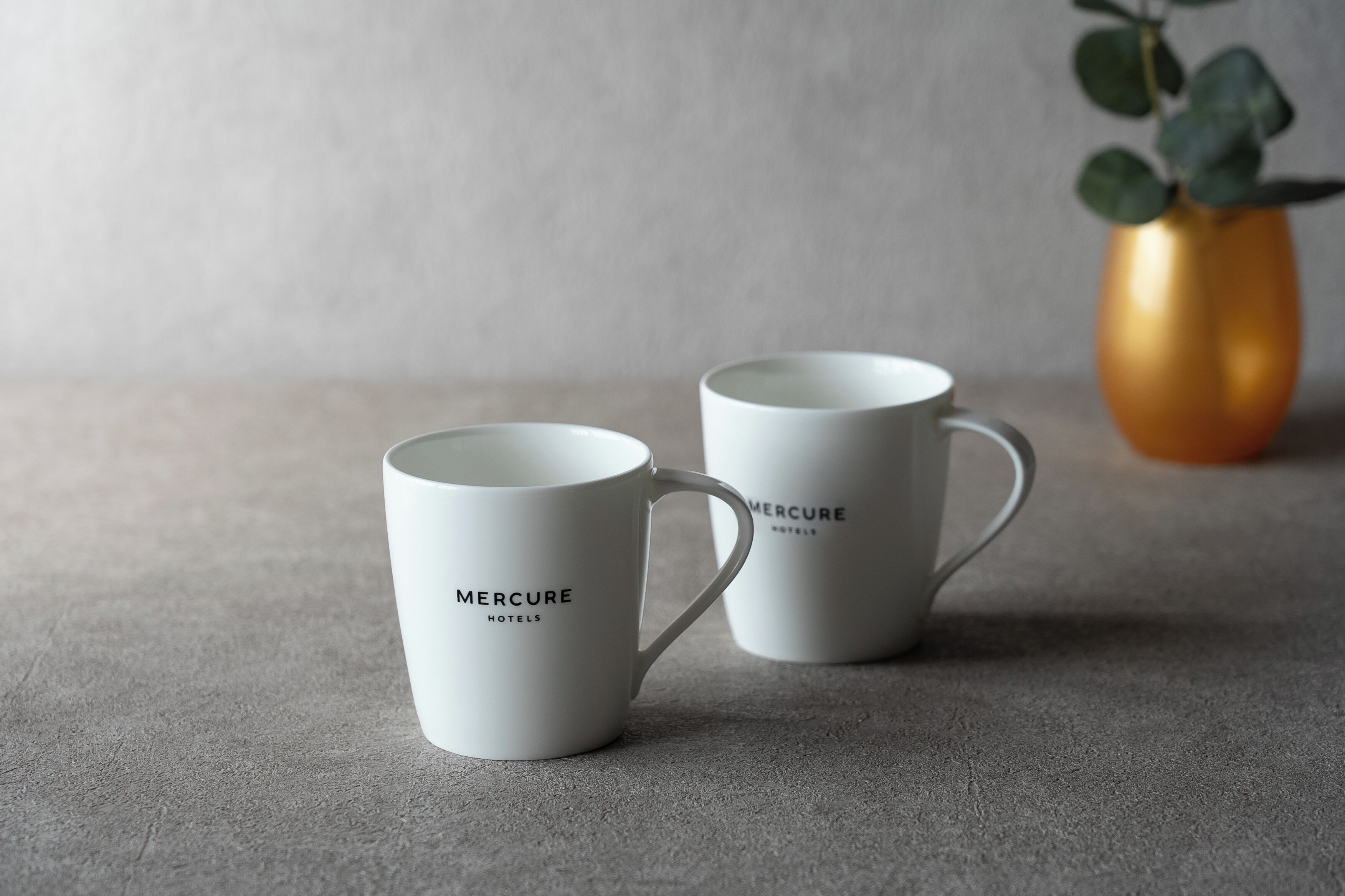 Two white mugs with "Mercure Hotels" text, placed on a textured surface with a blurred background.