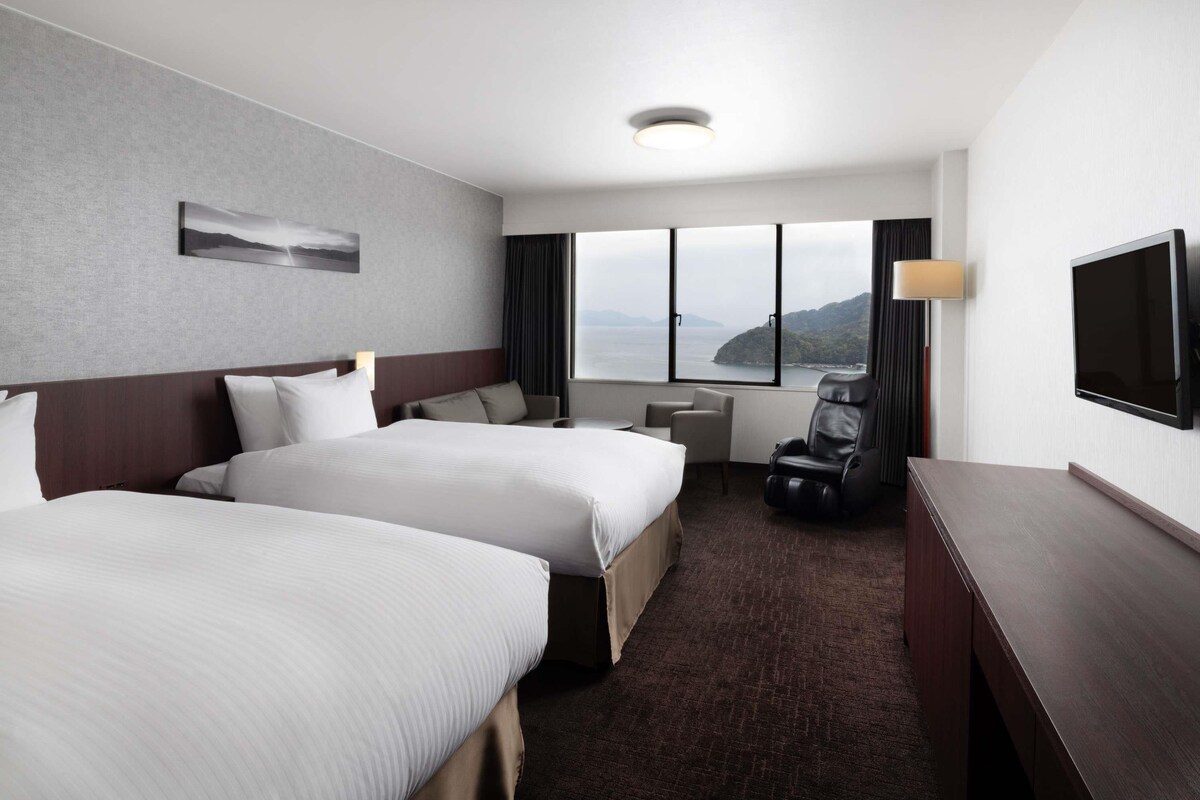 A hotel room with two beds, a desk, a chair, a TV, and a view of the sea.
