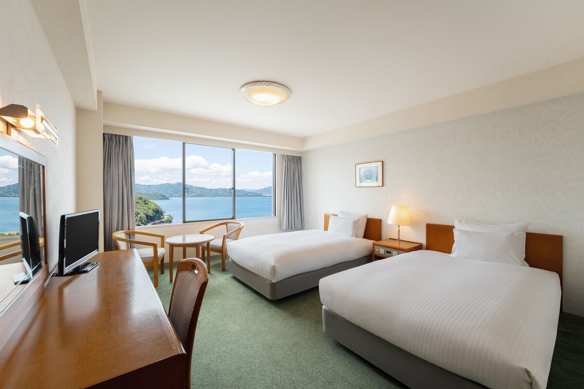 Ein Hotelzimmer mit zwei Betten, einem Schreibtisch mit Computer, einem Stuhl, einem Fenster mit Meerblick und einer Deckenleuchte.