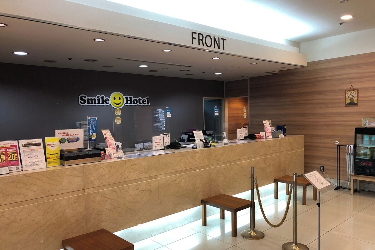 A hotel reception desk with a 'FRONT' sign above, a smiley face logo, and various informational posters. There are two wooden stools in front of the desk, and a Coca-Cola refrigerator to the right.