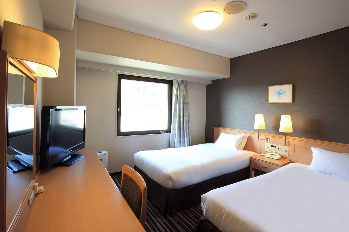 Ein Hotelzimmer mit zwei Betten, einem Schreibtisch mit Fernseher, einem Fenster mit Vorhängen und einer Wandlampe.