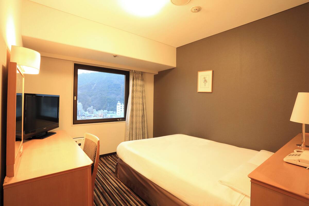 Ein Hotelzimmer mit einem Bett, einem Schreibtisch mit Fernseher, einem Sessel, einem Fenster mit Blick auf eine Stadtlandschaft und einem Wandbild.