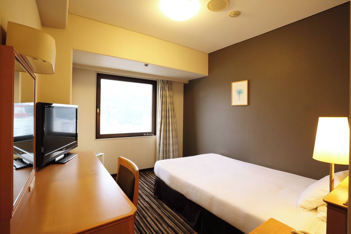 Ein Hotelzimmer mit einem Bett, einem Schreibtisch mit Fernseher, einem Sessel, einem Fenster mit Vorhängen und einem Wandbild.