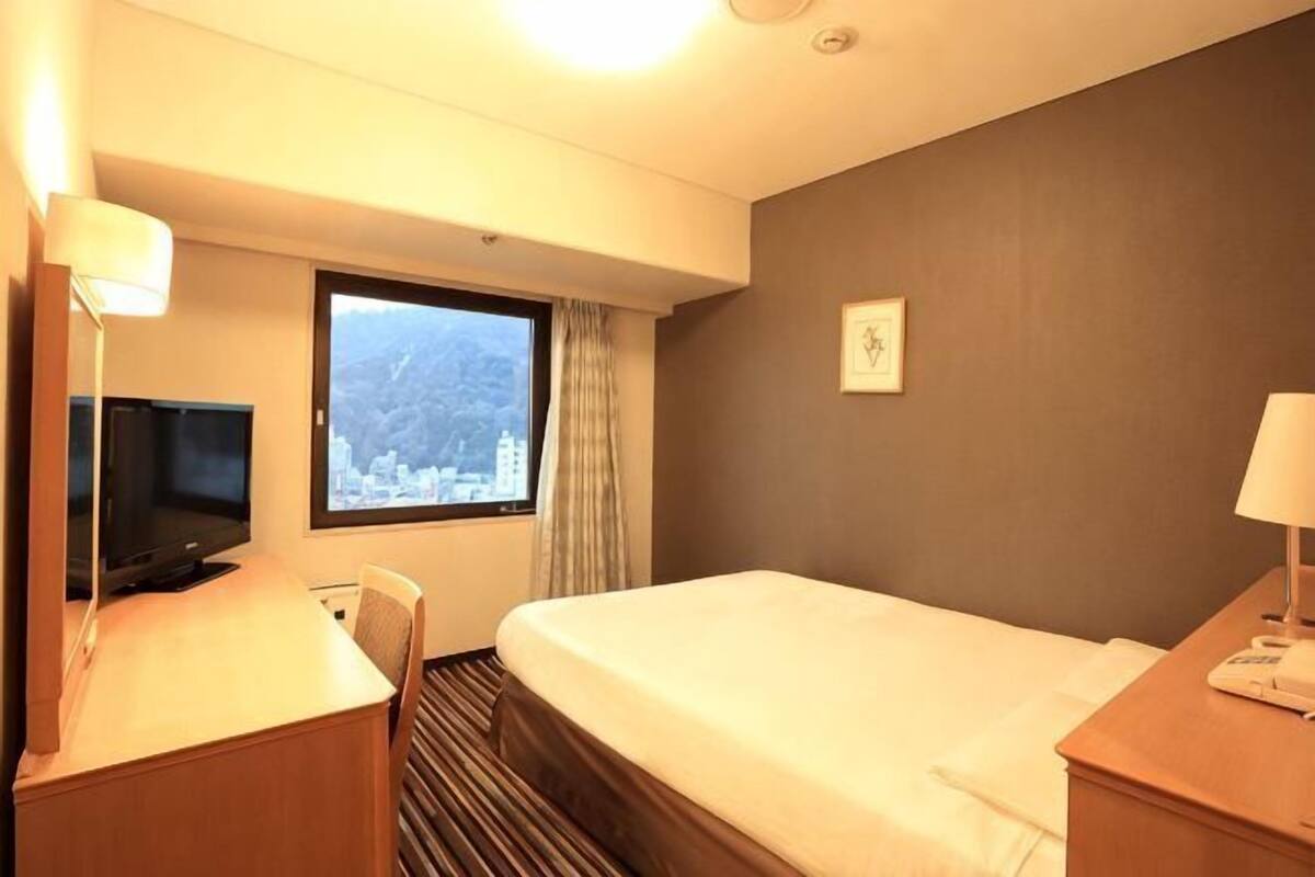 Ein Hotelzimmer mit Bett, Schreibtisch mit Fernseher, Fenster mit Blick auf die Stadt und einem an der Wand befestigten Telefon.