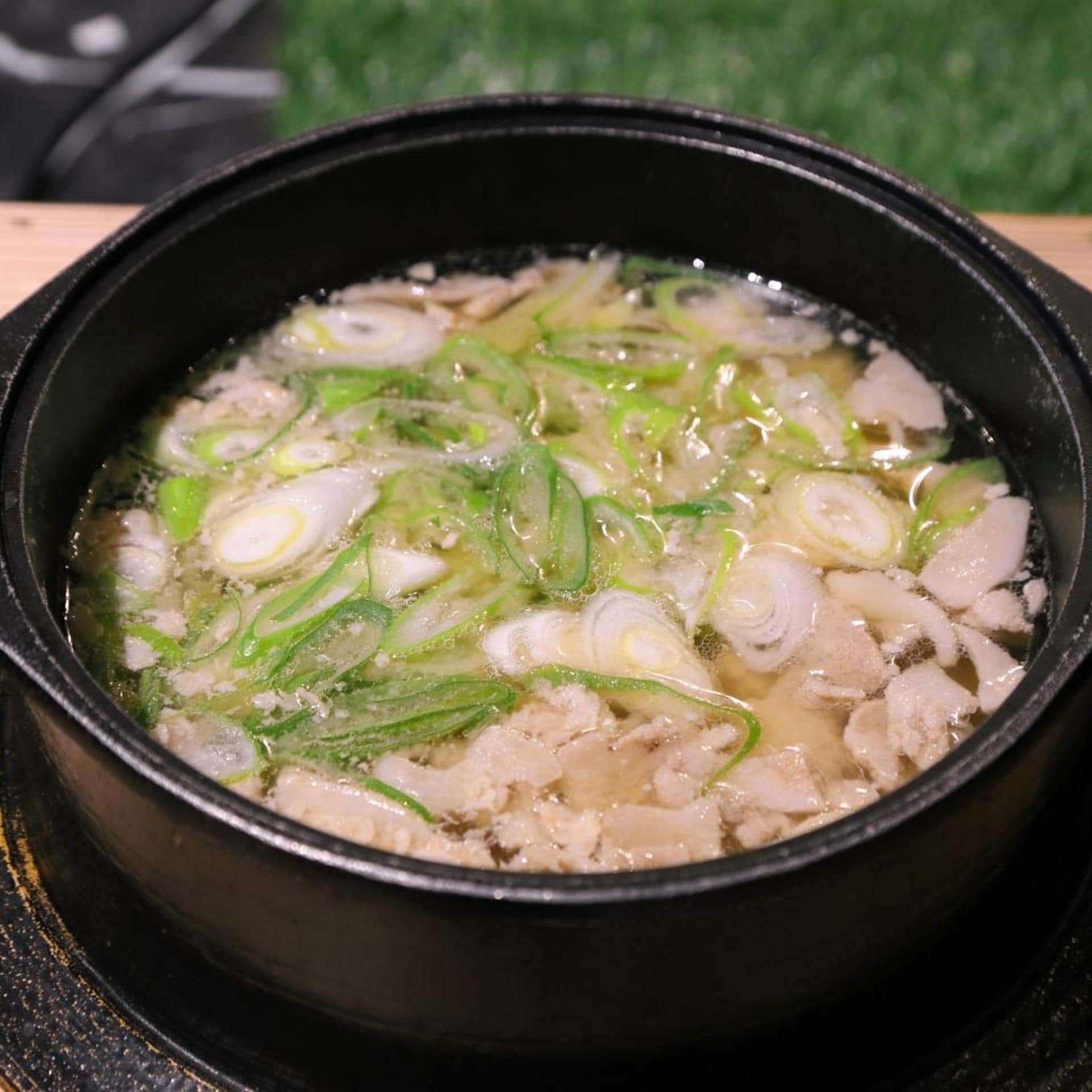A bowl of soup with sliced green onions and white chunks, possibly meat, in a black bowl on a table.