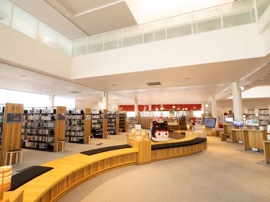 Ett rymligt bibliotek med många bokhyllor, en central läshörna och en stor övre våning med stora fönster.