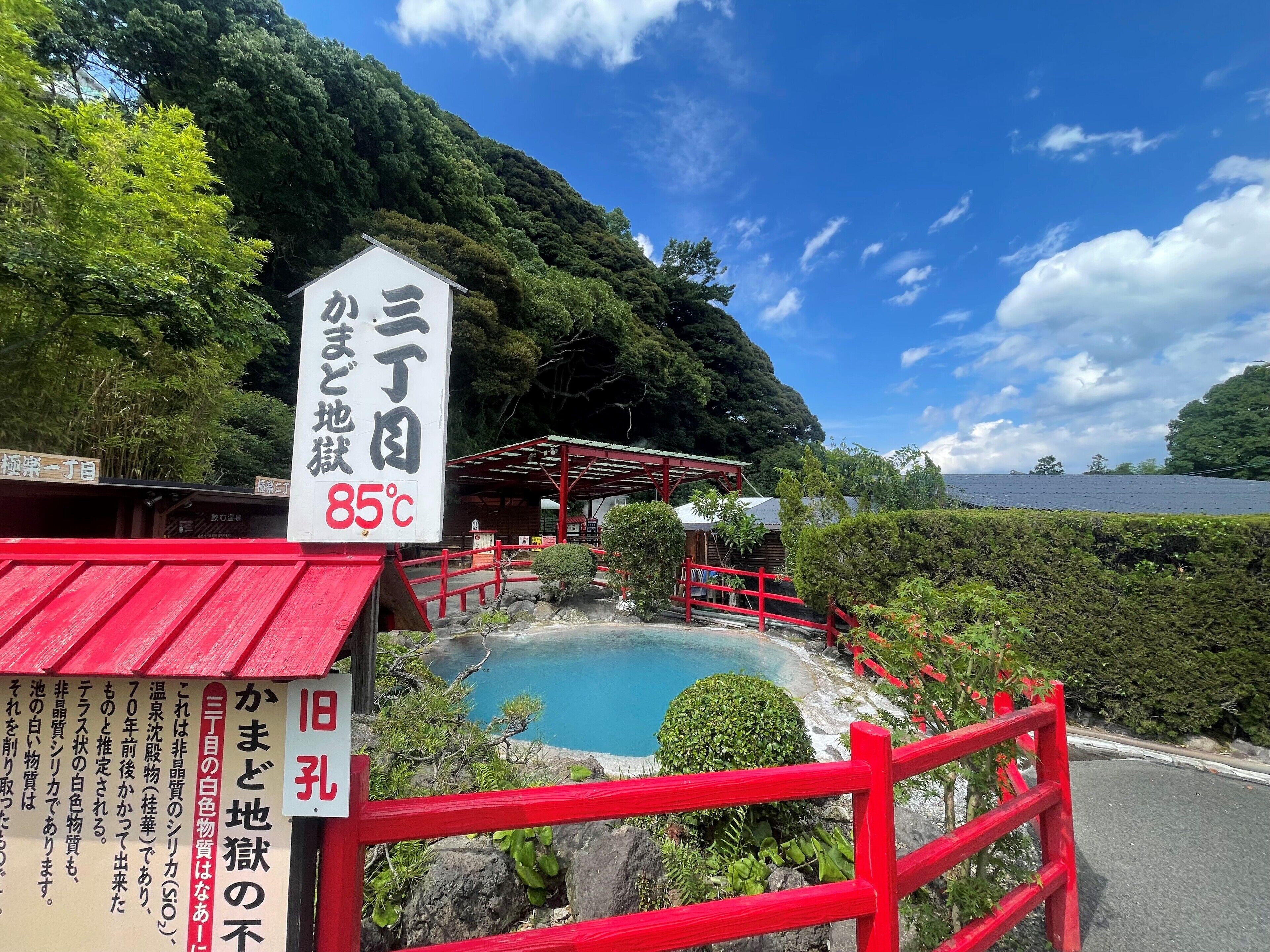 Un balneario japonés tradicional con valla de madera roja, rodeado de exuberante follaje y cielo azul despejado.