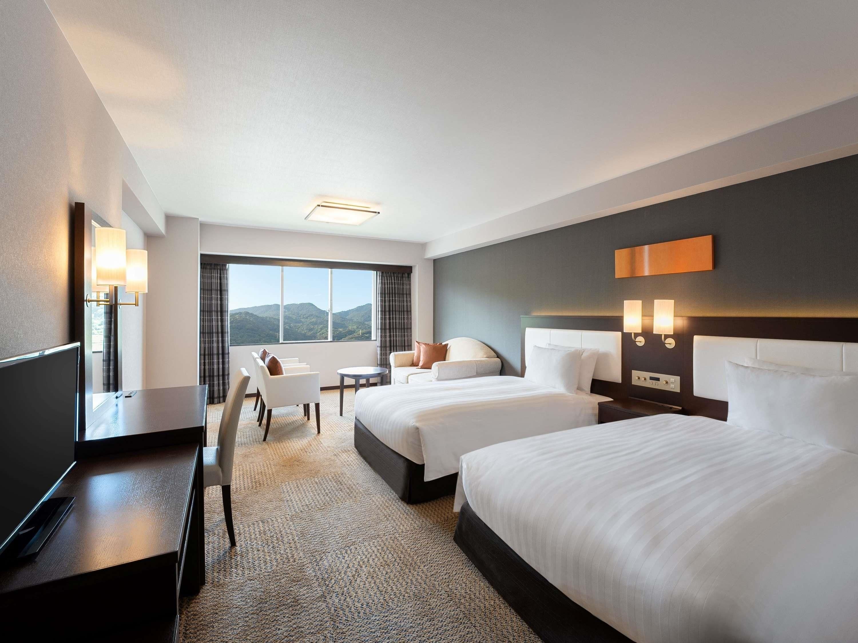 A hotel room with two beds, a desk, a chair, a small table, and a large window with a mountain view.