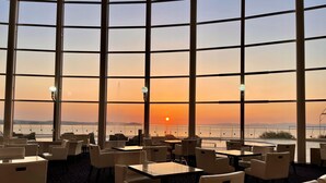 Un restaurante con grandes ventanales que ofrece vista al atardecer. Mesas y sillas dispuestas para comer.