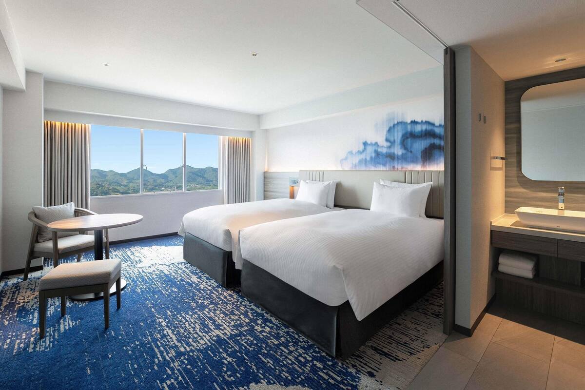 Een hotelkamer met twee bedden, een klein tafeltje, een stoel, een groot raam met uitzicht, en een badkamer met spiegel en wastafel.