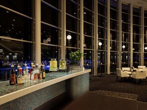 A bar area with a tiled counter, bottles, and glasses, large windows, and seating.