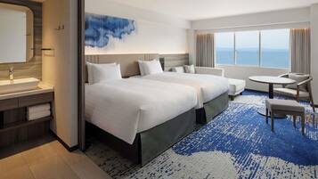 A hotel room with two beds, a desk, a chair, and a view of the sea.